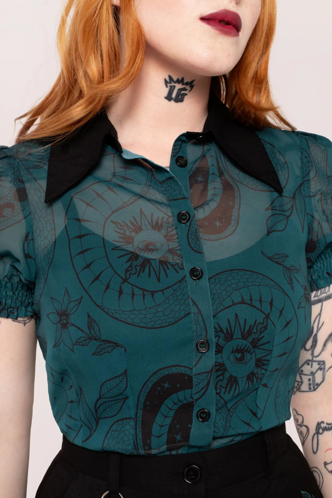 Person wearing a teal blouse with black collar and black buttons, featuring abstract designs.