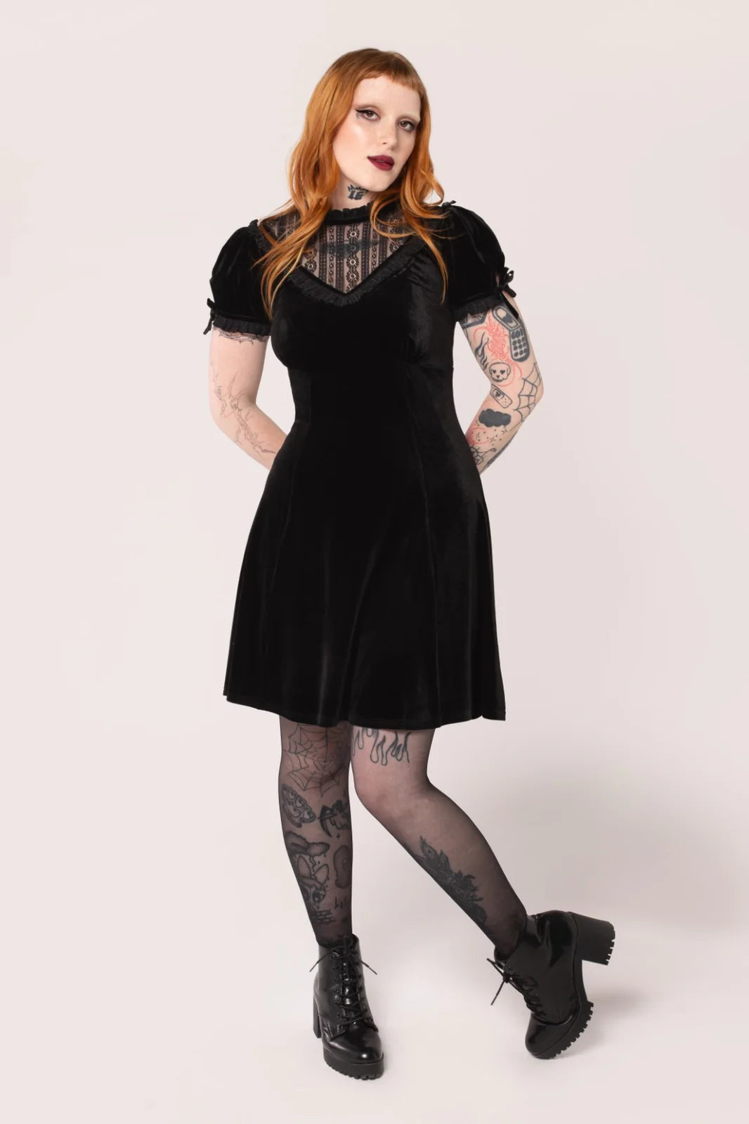 A person wearing a black velvet knee-length dress with a high neckline, black striped lace panel on chest, and short puff sleeves. The dress has a flared skirt and is accessorized with a black top hat, black boots, and red lipstick.