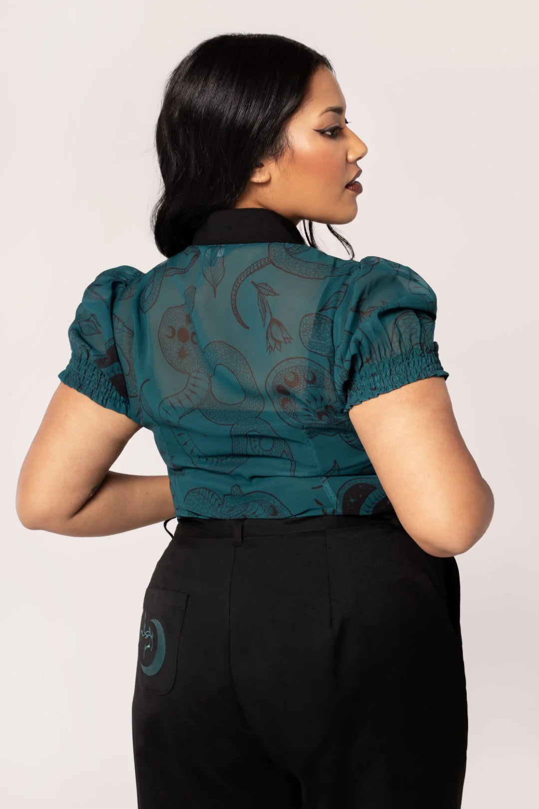 Woman wearing a teal blouse with short sleeves and black pants on a light pink background