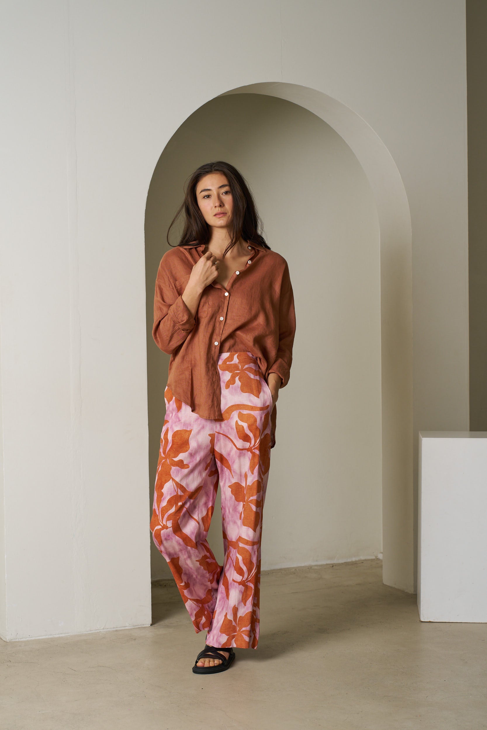 Woman wearing a rust-colored shirt and pink floral pants standing in an archway.